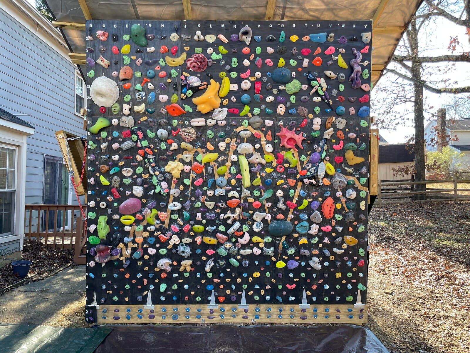 A steep black climbing wall covered in colorful holds, under a roof in a backyard