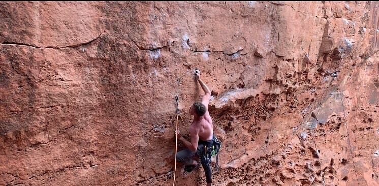 A shirtless climber on lead on a red sandstone wall, rope clipped to a quickdraw