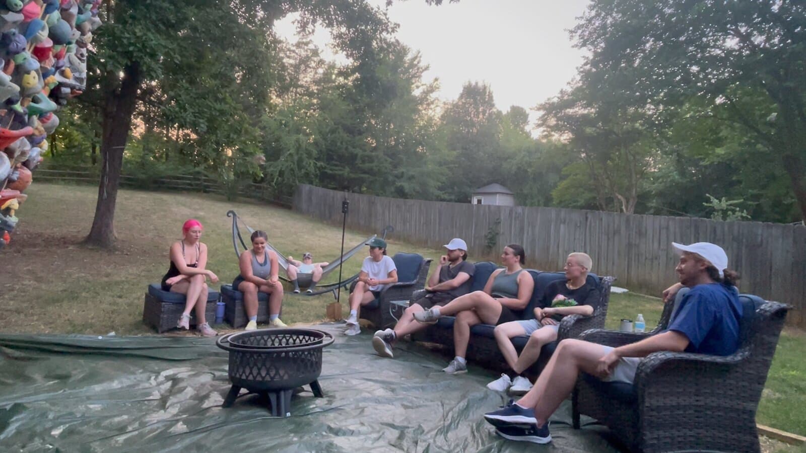 Contestants seated around a fire pit in a backyard at dusk