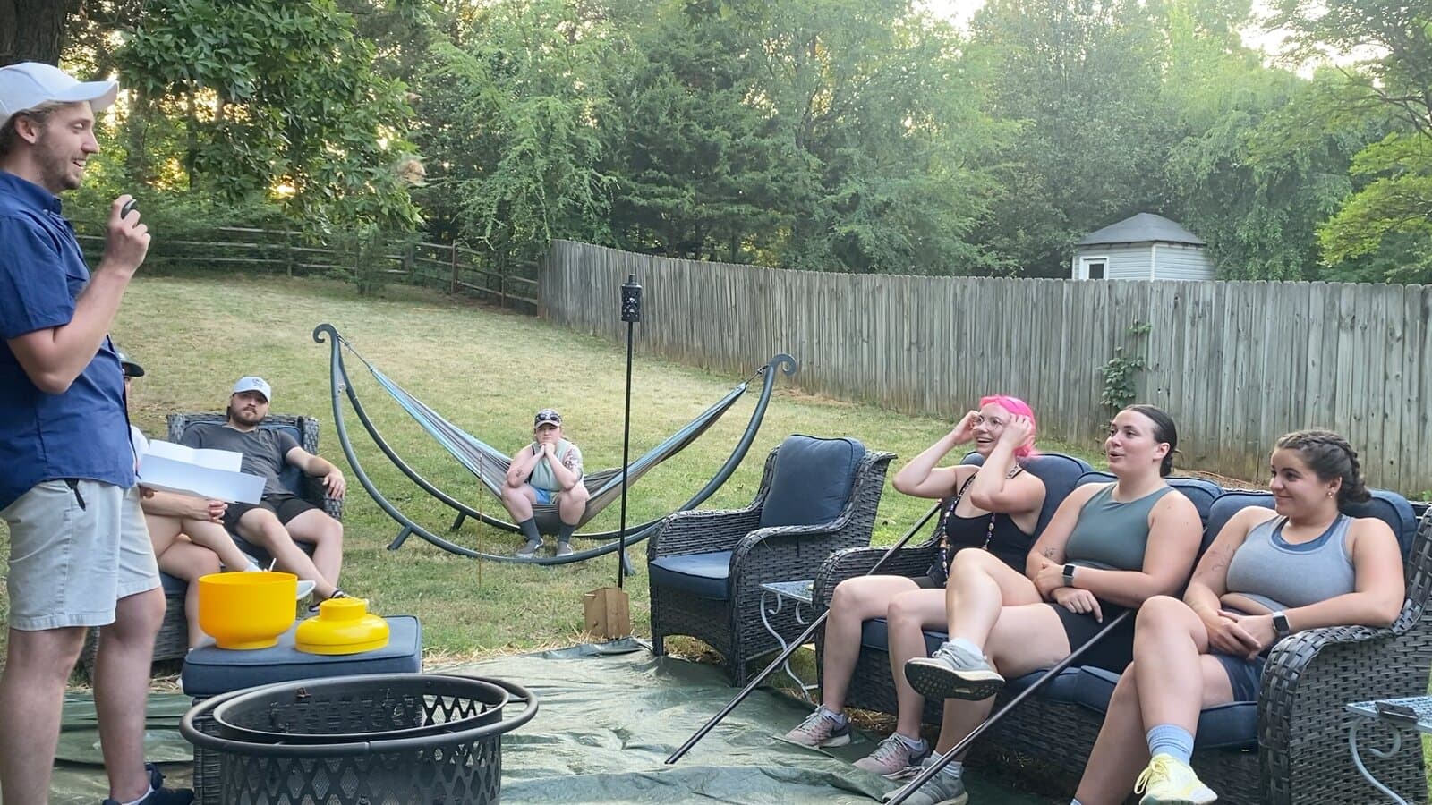 The host standing with papers beside the yellow urn while contestants sit on outdoor couches